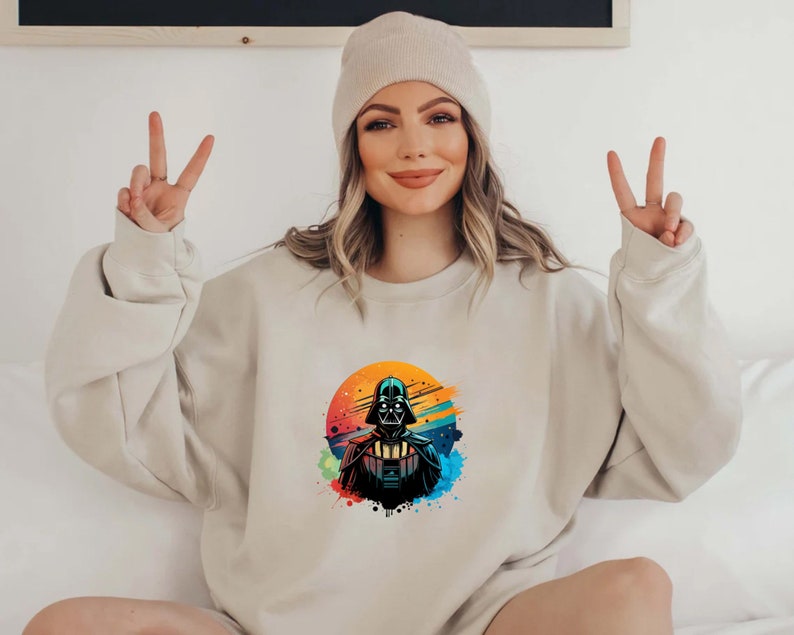 Op de afbeelding: Beige sweatshirt met een kleurrijke Darth Vader-afbeelding. Het ontwerp heeft een levendige zonsondergangachtige achtergrond met kleurspatten. De sweatshirt heeft lange mouwen en een ronde hals. Een beige muts is ook zichtbaar.