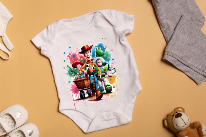 Op de afbeelding: Witte baby romper met een kleurrijk aquarelontwerp met Woody, Buzz Lightyear en Jessie uit Toy Story. De personages staan voor een kar met kleurrijke verfspatten op de achtergrond.