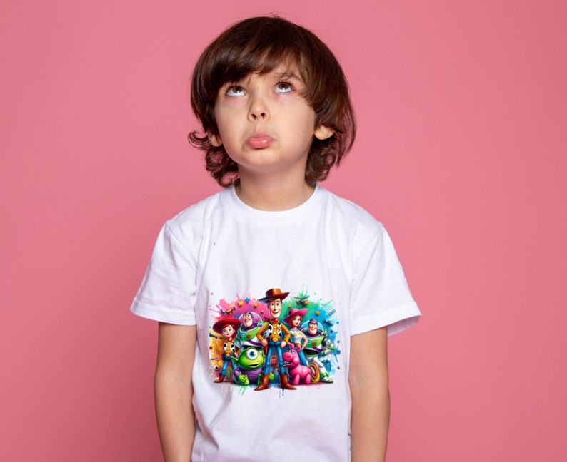 Op de afbeelding: Een wit T-shirt met een kleurrijke grafische print van personages uit de Toy Story-filmfranchise. De personages zijn onder andere Woody, Buzz Lightyear, Jessie en anderen. De grafiek is omgeven door kleurrijke verfspatten.