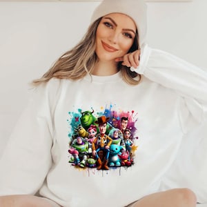 Op de afbeelding: Een wit sweatshirt met een kleurrijke grafische print van personages uit de Toy Story-franchise. De personages zijn Woody, Buzz Lightyear, Jessie en Rex. De grafiek is omringd door kleurrijke verfspatten.