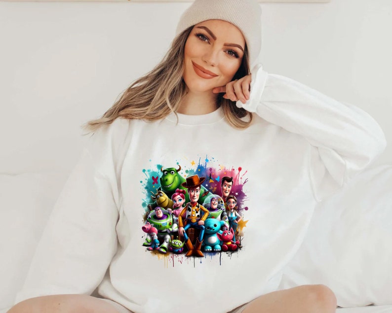 Op de afbeelding: Een wit sweatshirt met een kleurrijke grafische print van personages uit de Toy Story-franchise. De personages zijn Woody, Buzz Lightyear, Jessie en Rex. De grafiek is omringd door kleurrijke verfspatten.