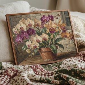 以下が含まれることがあります： テラコッタの鉢植えに入った蘭の額入り絵画。花はクリーム色、紫、オレンジ色で、レンガの壁を背景にしています。絵画は木製のフレームに入っており、模様入りのキルトの上に置かれています。