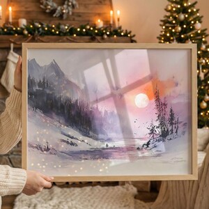 Op de afbeelding: Een ingelijste aquarel van een winterlandschap. Het kunstwerk toont besneeuwde bergen, een bos en een meer onder een zonsondergang met een grote, heldere maan. Het frame is gemaakt van lichtgekleurd hout.