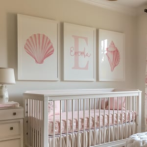 May include: A nursery with a white crib, dresser, and armchair. Pink seashell art hangs above the crib, with the name "Emma" displayed. The room features a floral rug, a tufted ottoman, and a lamp.