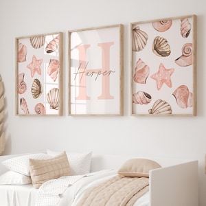 May include: Three framed wall art pieces. Two feature seashell and starfish illustrations in shades of peach and brown. The center frame displays the name "Harper" with a large pink letter "H". The room has a white bed with a beige quilt.
