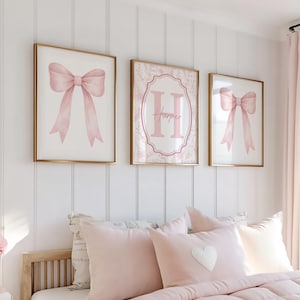 Op de afbeelding: Drie ingelijste wandkunstwerken met gouden frames. Twee tonen roze aquarel strikken. Het middelste stuk heeft de naam "Harper" in roze. Het bed eronder heeft roze kussens en een dekbed.
