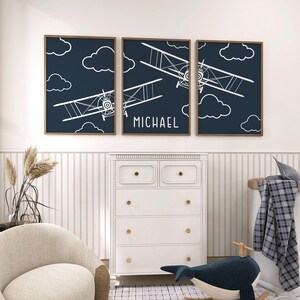 May include: Three framed prints with a navy blue background and white silhouettes of aeroplanes flying in a cloudy sky. The middle print has the name "MICHAEL" written below the aeroplanes.