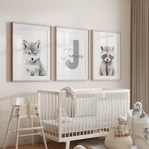 May include: Three framed prints for a nursery, featuring a wolf, a raccoon, and a letter J with the name "James" written below. The prints are hung on a white wall above a white crib.