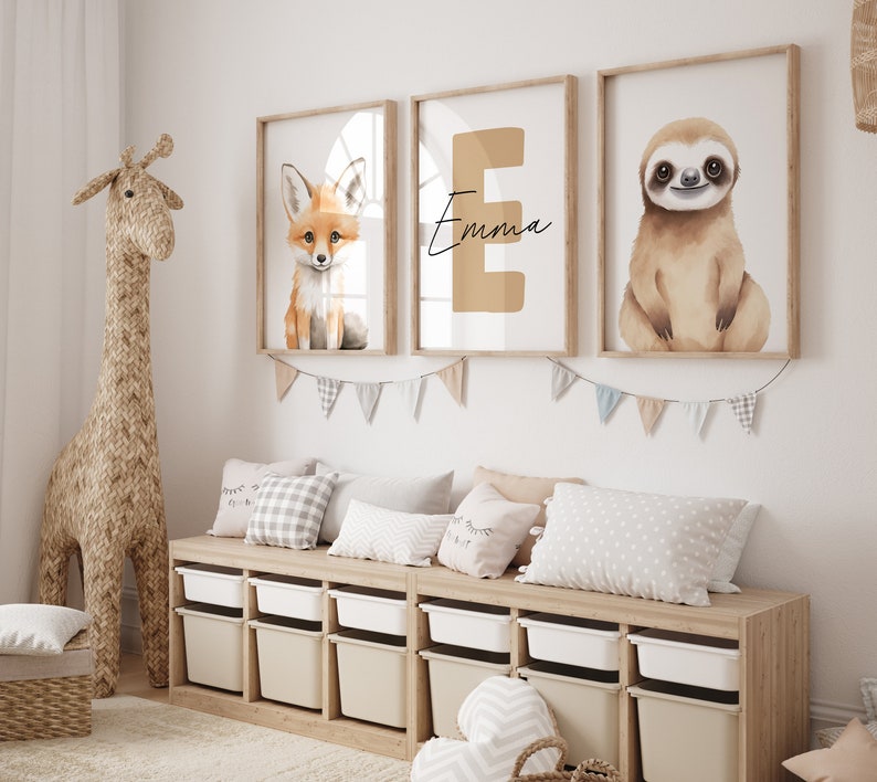 May include: Three framed prints with watercolor illustrations of a fox, a sloth, and the letter E with the name "Emma" written below. The prints are hanging on a white wall above a wooden shelf with storage bins.