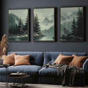 May include: Three framed landscape art prints showing misty mountain scenes with evergreen trees and a lake. The artwork is displayed above a blue sofa with orange cushions and a dark grey throw. A modern living room setting.