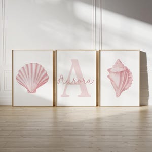 May include: Three framed art prints with a coastal theme. The prints feature a pink seashell, the letter "A" with the name "Aurora", and a conch shell. The artwork is set against a white background with a light wood frame.