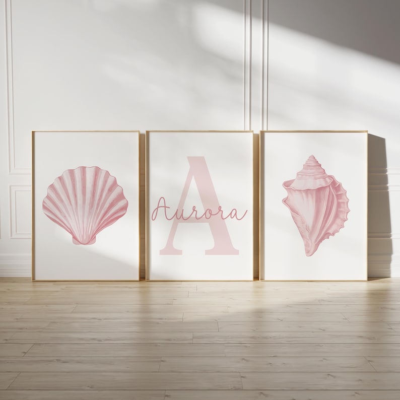 May include: Three framed art prints with a coastal theme. The prints feature a pink seashell, the letter "A" with the name "Aurora", and a conch shell. The artwork is set against a white background with a light wood frame.