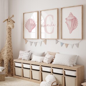 May include: Three framed prints with seashell illustrations and a personalized name. The prints are in a light pink color and are displayed above a wooden storage unit with white bins. A woven giraffe and pillows complete the room's decor.