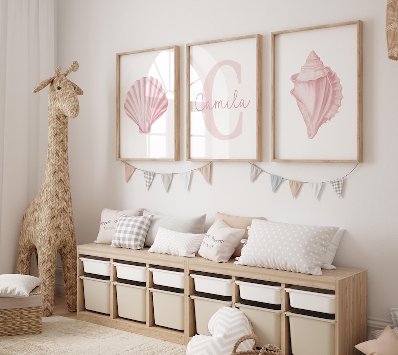 May include: Three framed prints with seashell illustrations and a personalized name. The prints are in a light pink color and are displayed above a wooden storage unit with white bins. A woven giraffe and pillows complete the room's decor.