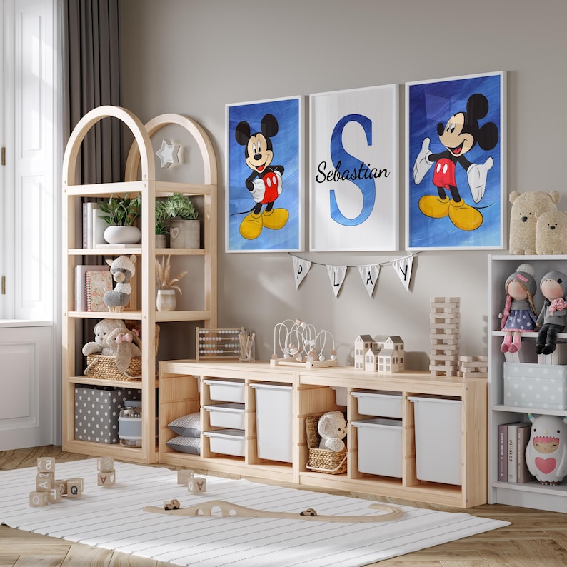 Op de afbeelding: Een lichte houten kinderkamer met een houten boekenkast, een houten speelgoedopbergkast en drie ingelijste prints met Mickey Mouse. De prints zijn op een lichtblauwe achtergrond met witte lijsten. De middelste print heeft de letter "S" in blauw met de naam "Sebastian" eronder. De speelkamer heeft een lichtgrijs tapijt met een houten treinspoor.