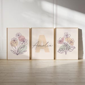以下が含まれることがあります： 花柄のデザインと名前「アメリア」が書かれた、ベージュの額縁に入ったプリントが3枚。花柄は紫、ピンク、緑の色合いで描かれています。