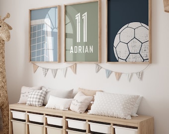 Puede incluir: Tres impresiones enmarcadas en una pared blanca sobre un banco de madera con cojines. Las impresiones presentan un diseño geométrico azul y blanco, un fondo verde con el número 11 y el nombre Adrian, y un fondo azul con un balón de fútbol blanco.