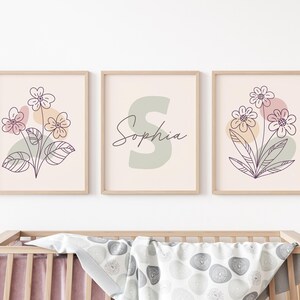 以下が含まれることがあります： 花柄と「ソフィア」という名前が描かれた、ミニマルなデザインの額装されたプリント3枚。プリントは薄いピンク色の背景に、桃色、緑色、紫色の抽象的な形が描かれています。