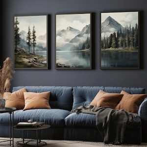 May include: Three framed landscape art prints depicting mountain and lake scenes. The artwork features muted colors of blues, greens, and grays. The prints are displayed above a blue sofa with orange pillows and a gray throw blanket.