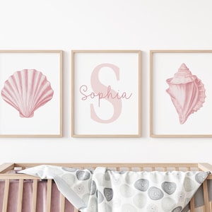 May include: Three framed prints featuring pink seashell illustrations and the name "Sophia". The prints are in light wood frames. The artwork is displayed above a wooden crib with a patterned blanket.