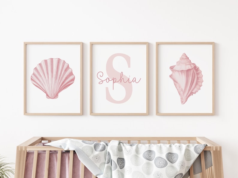 May include: Three framed prints featuring pink seashell illustrations and the name "Sophia". The prints are in light wood frames. The artwork is displayed above a wooden crib with a patterned blanket.