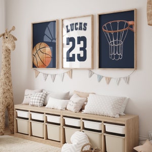 Può includere: Tre stampe incorniciate a tema basket. La prima stampa presenta una palla da basket su sfondo blu. La seconda stampa ha uno sfondo bianco con il nome Lucas e il numero 23 in blu. La terza stampa mostra un canestro da basket su sfondo blu.