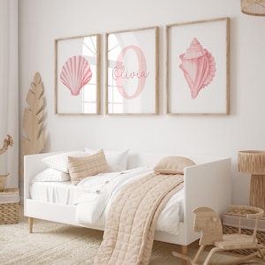 May include: Three framed wall art prints with a seashell, the letter "O" with the name "Olivia", and a conch shell in pink hues. The prints are in light wood frames. The room has a white bed with a beige quilt and pillows.