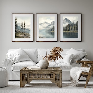 May include: Three framed landscape art prints depicting serene mountain lake scenes. Each print features a different view of mountains, water, and trees, with a soft, watercolor-like aesthetic. The frames are a warm brown, complementing the natural tones of the artwork.