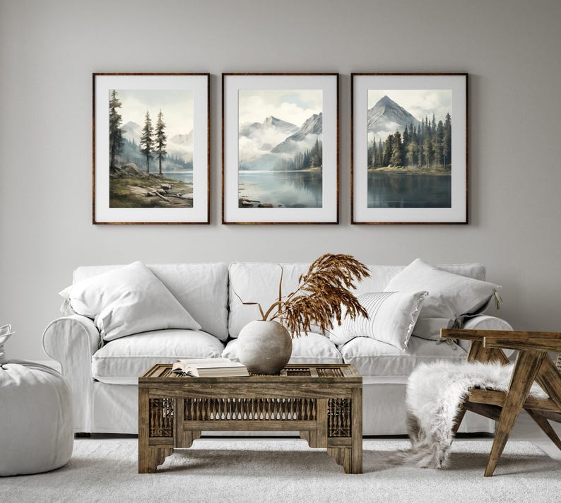 May include: Three framed landscape art prints depicting serene mountain lake scenes. Each print features a different view of mountains, water, and trees, with a soft, watercolor-like aesthetic. The frames are a warm brown, complementing the natural tones of the artwork.
