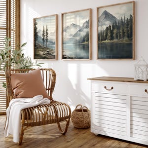 May include: Three framed landscape art prints depicting mountain and lake scenes in muted tones of blue, green, and brown. The prints are displayed above a white cabinet and a wicker chair with a pillow and blanket.