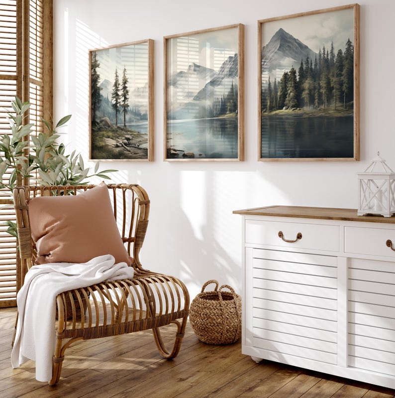 May include: Three framed landscape art prints depicting mountain and lake scenes in muted tones of blue, green, and brown. The prints are displayed above a white cabinet and a wicker chair with a pillow and blanket.