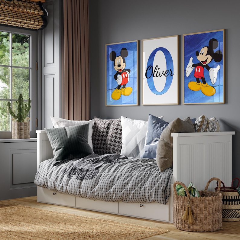 Op de afbeelding: Drie ingelijste prints met Mickey Mouse op een blauwe achtergrond. De middelste print heeft de naam "Oliver" in een witte cirkel op een blauwe achtergrond. De prints hangen aan een grijze muur boven een wit dagbed met kussens en een deken.