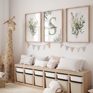 May include: Three framed prints with a green and white floral design. The middle print features the name "Sophia" in a green circle. The prints are hanging on a white wall above a wooden shelf with storage bins and pillows.