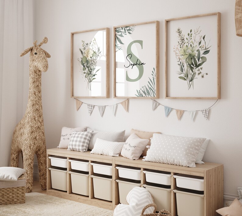 May include: Three framed prints with a green and white floral design. The middle print features the name "Sophia" in a green circle. The prints are hanging on a white wall above a wooden shelf with storage bins and pillows.