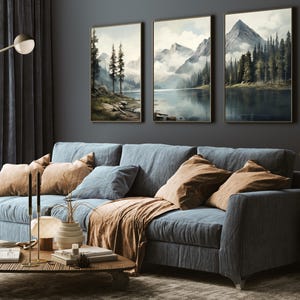 May include: A living room scene featuring a blue sofa with tan and blue pillows and a brown throw blanket. Above the sofa are three framed landscape paintings depicting mountains and lakes. A wooden coffee table with decor sits in front of the sofa.