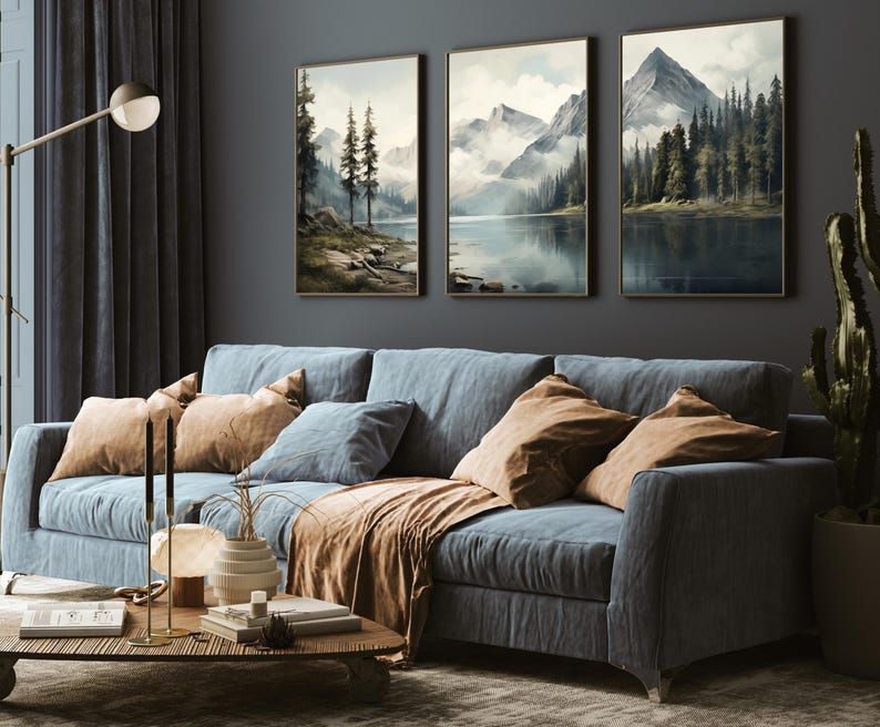 May include: A living room scene featuring a blue sofa with tan and blue pillows and a brown throw blanket. Above the sofa are three framed landscape paintings depicting mountains and lakes. A wooden coffee table with decor sits in front of the sofa.