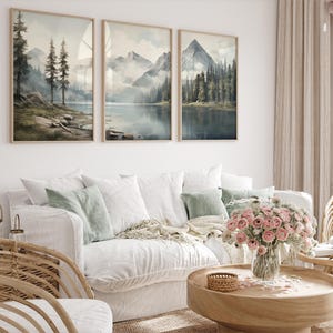 May include: Three framed landscape art prints depicting mountain and lake scenes in muted tones of blue, green, and gray. The artwork hangs above a white sofa with decorative pillows and a wooden coffee table with a flower arrangement.