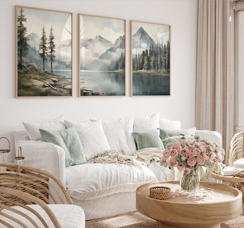 May include: Three framed landscape art prints depicting mountain and lake scenes in muted tones of blue, green, and gray. The artwork hangs above a white sofa with decorative pillows and a wooden coffee table with a flower arrangement.