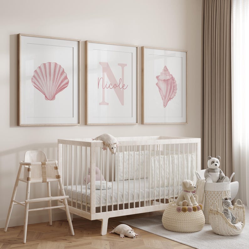May include: Three framed prints with seashell and initial designs in pink hues hang above a white crib. The middle print features the name "Nicole". A high chair, baskets with stuffed animals, and toy mice complete the nursery decor.