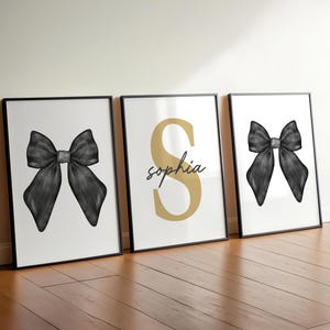 May include: Three framed art prints leaning against a wall. Two prints feature a black bow on a white background. The center print has a gold letter "S" and the name "Sophia" in script.