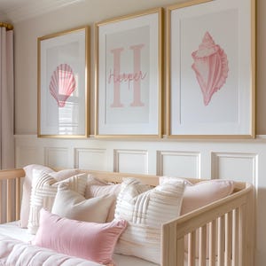 May include: Three framed prints with gold frames hang above a crib. The prints feature pink seashell art and the name "Harper" in pink. The crib is filled with pink and cream pillows and a pink blanket. The room has a soft, feminine aesthetic.