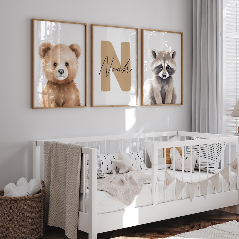 May include: Three framed prints with a brown bear, the letter N with the name Noah written in cursive, and a raccoon. The prints are hanging on a white wall above a white crib with a white blanket and pillows.