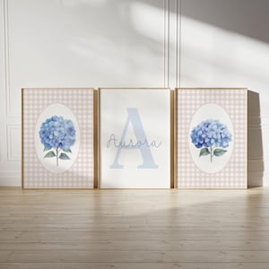Pode incluir: Três peças de arte de parede emolduradas. Duas apresentam uma hortênsia azul em uma moldura oval em um fundo xadrez rosa e branco. A peça central tem uma grande letra "A" azul e o nome "Aurora" em itálico.
