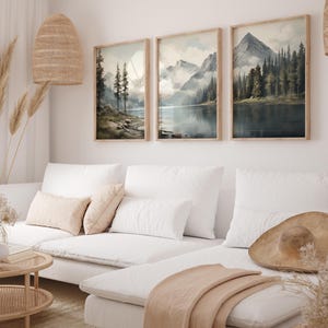 May include: Three framed landscape art prints depicting mountain scenes with lakes and forests. The prints have light brown wooden frames and are displayed above a white sectional sofa. The artwork features muted colors and a serene, natural theme.