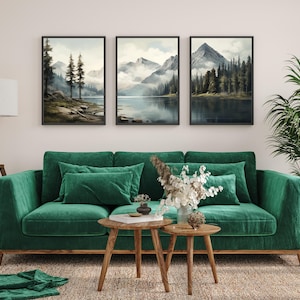 May include: Three framed landscape art prints depicting serene mountain and lake scenes. The artwork features muted colors and a tranquil atmosphere. The prints are displayed above a green velvet sofa with matching pillows, complemented by wooden side tables and a rug.