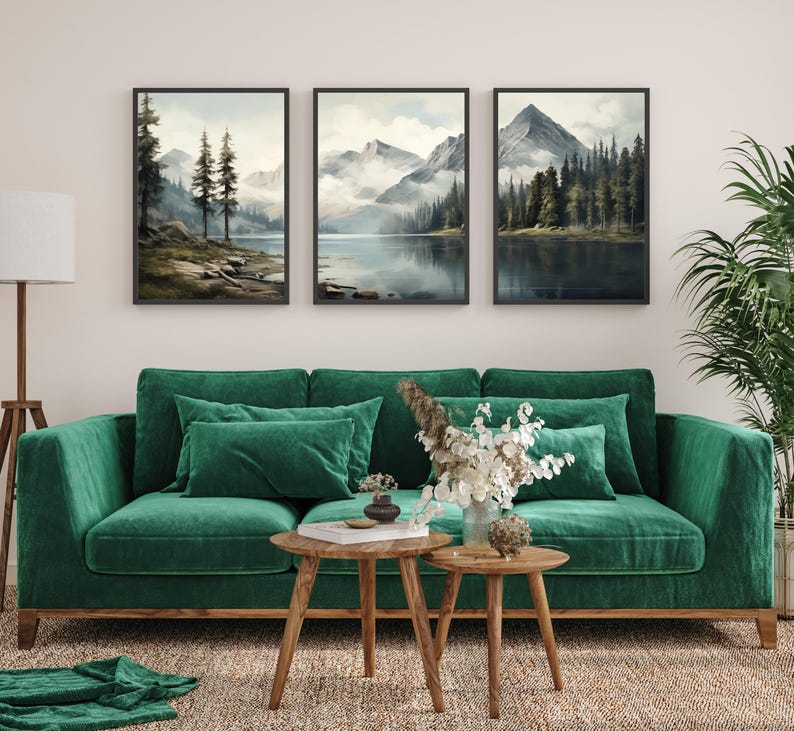 May include: Three framed landscape art prints depicting serene mountain and lake scenes. The artwork features muted colors and a tranquil atmosphere. The prints are displayed above a green velvet sofa with matching pillows, complemented by wooden side tables and a rug.