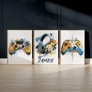 Puede incluir: Tres impresiones artísticas enmarcadas con temas de videojuegos. Las impresiones muestran un mando de juego amarillo y azul, unos auriculares y otro mando. Cada imagen tiene un efecto acuarela con salpicaduras de pintura y un marco dorado.