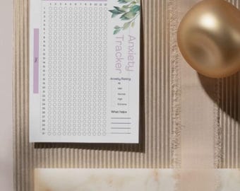 May include: Anxiety Tracker journal with a floral design. The white paper has a grid for tracking daily anxiety levels, with the words "Anxiety Tracker" and "What helps" printed on the page. The journal is on a textured surface.