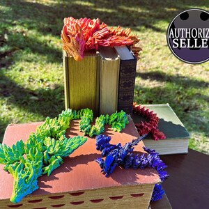 May include: A display of colorful, articulated dragon figurines in red, green, and blue, arranged on and around vintage books. The dragons are posed as if interacting with the books. The image includes an "AUTHORIZED SELLER" badge.