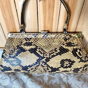 May include: Vintage handbag with a snakeskin pattern in tan, black, and blue. The bag has a structured rectangular shape with a silver-tone clasp and a curved handle. A decorative floral embellishment adorns the handle.